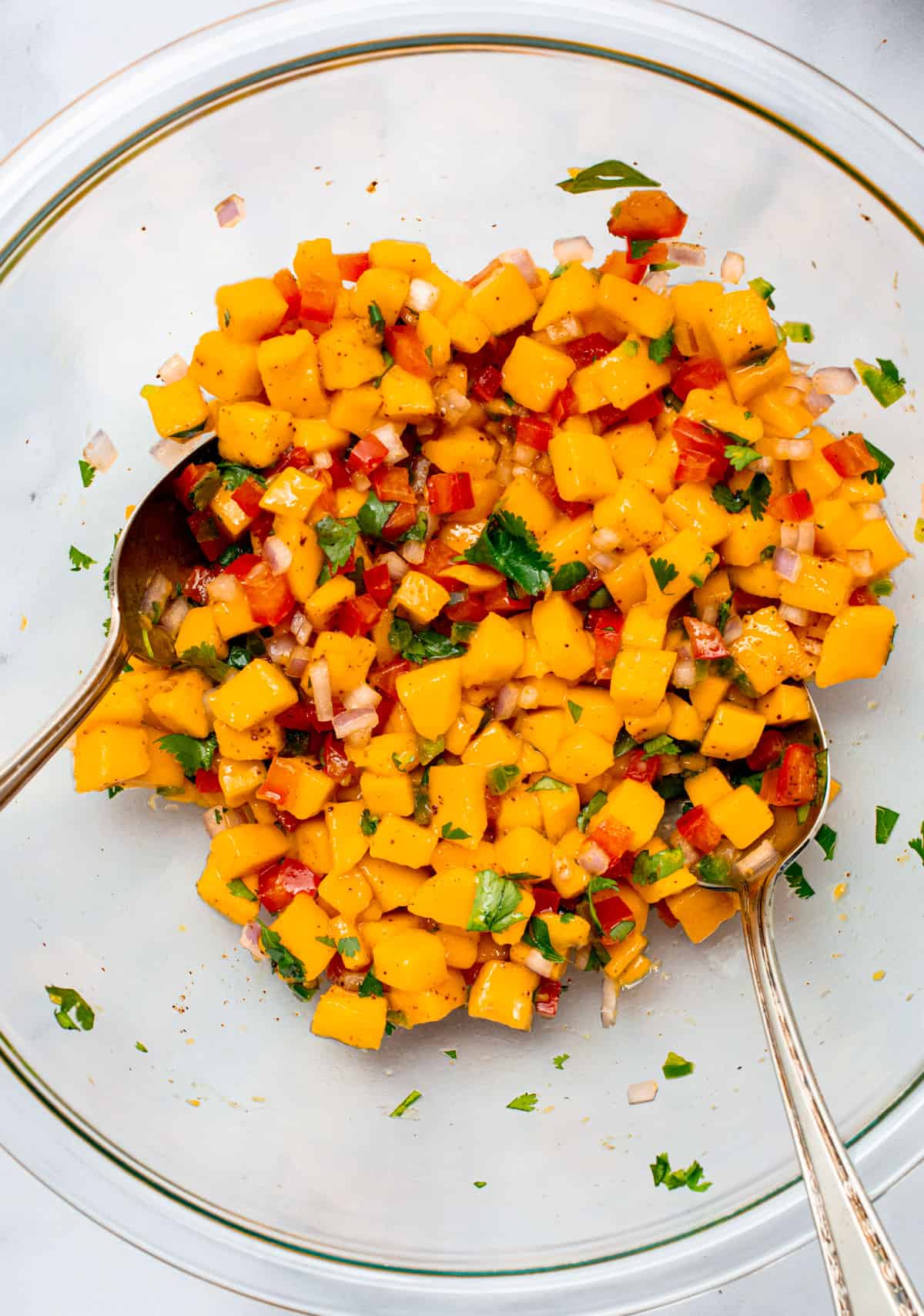 Freshly mixed mango salsa in a glass bowl with 2 mixing spoons.
