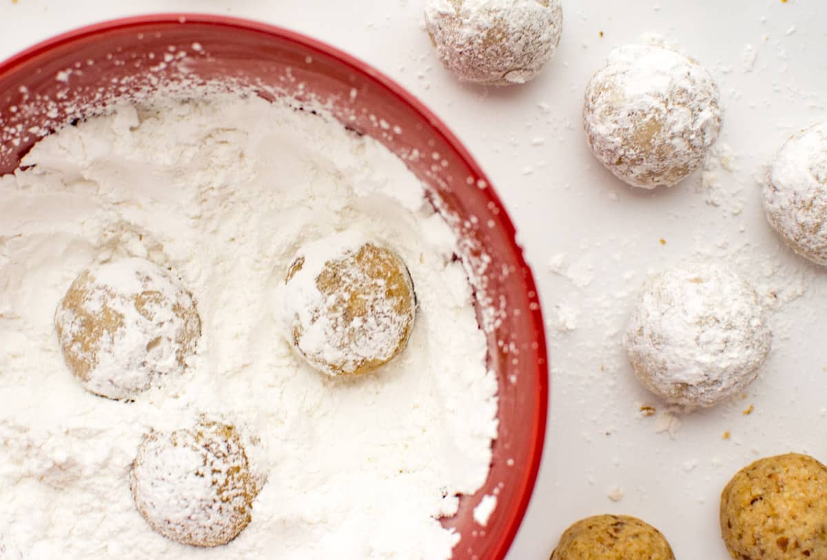 Vegan snowball cookies being rolled in a bowl of powdered sugar.