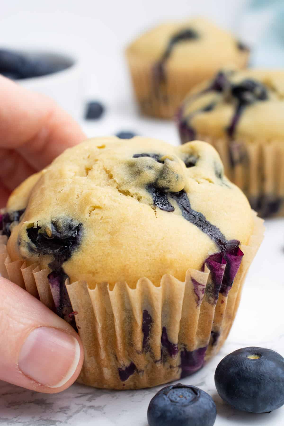 A hand grabbing a vegan blueberry muffin with more muffins and blueberries behind.