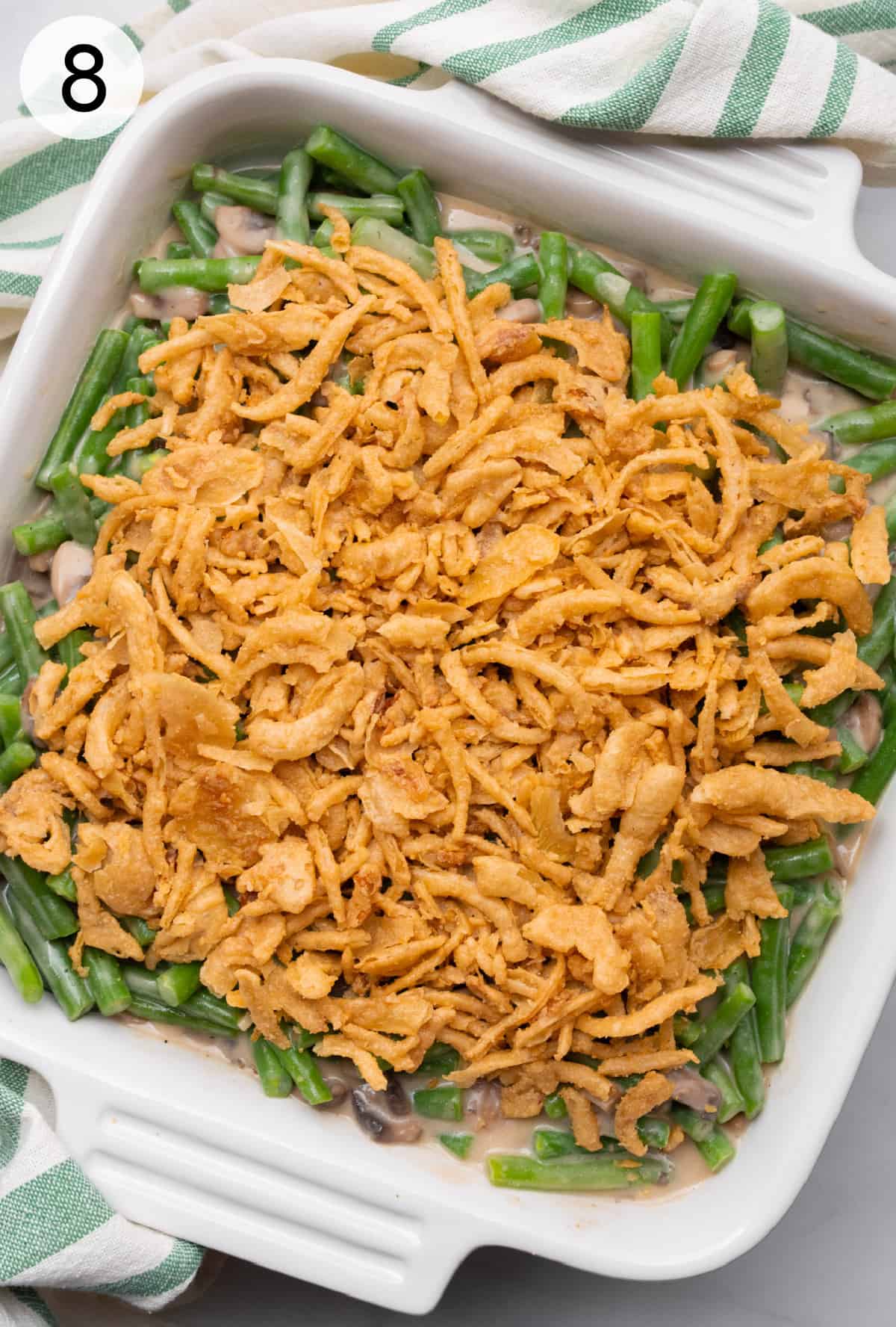 Vegan green bean casserole topped with french fried onions in a white square ceramic dish, before baking.