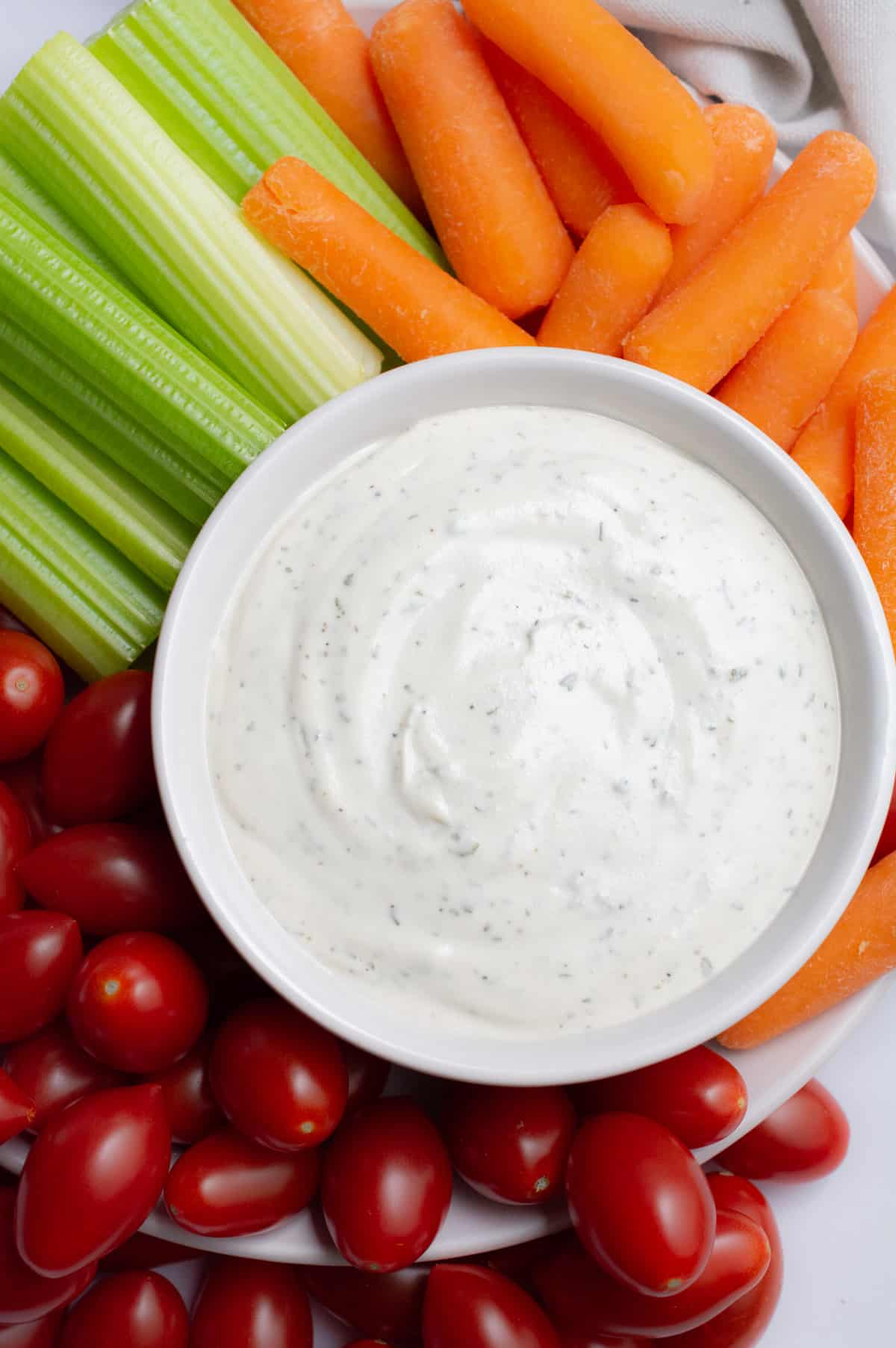 A bowl of vegan ranch dip on a plate with carrots, celery, and grape tomatoes.