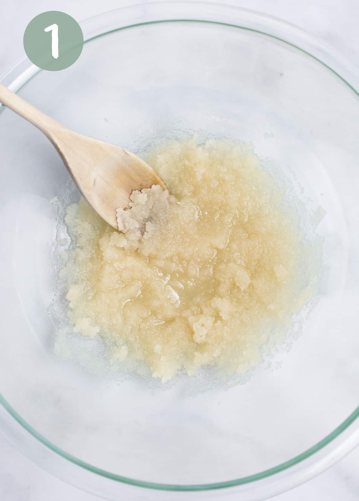 Oil and sugar mixed together with a wooden spoon in a glass bowl.