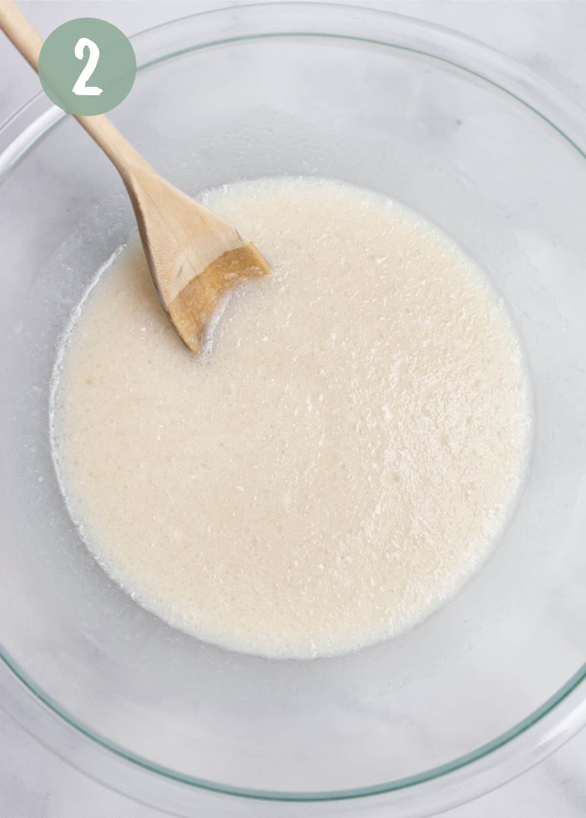 Oil, sugar, almond milk, yogurt and almond extract mixed with a wooden spoon in a glass bowl.