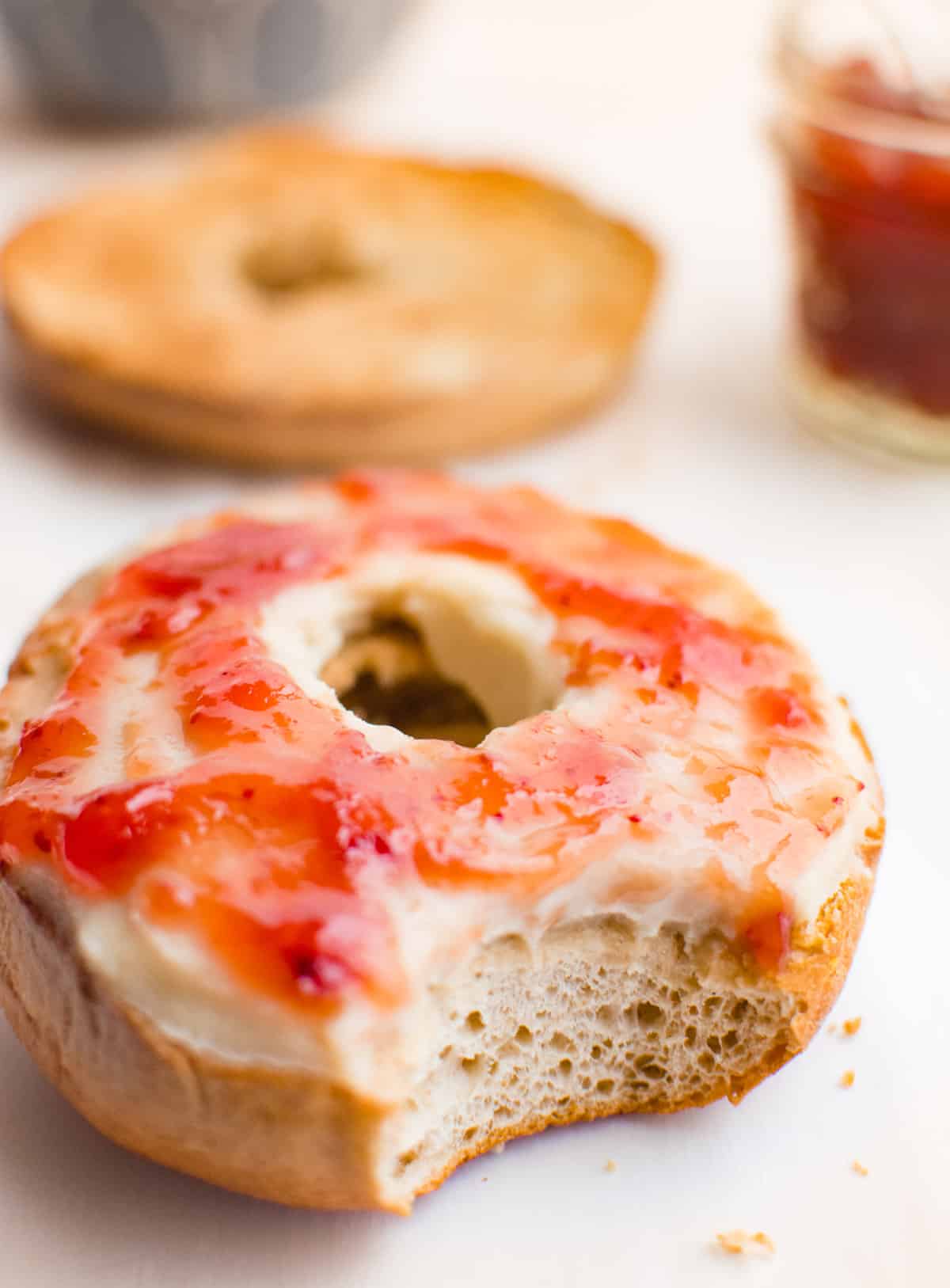 A plain bagel topped with cashew cream cheese and strawberry jam, with a bite taken out.