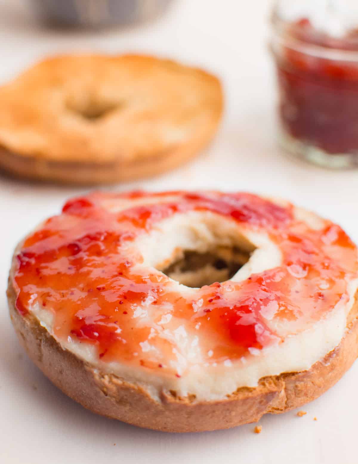 A toasted bagel spread with cashew cream cheese and strawberry preserves. There is a jar of preserves and other half of toasted bagel in the background.