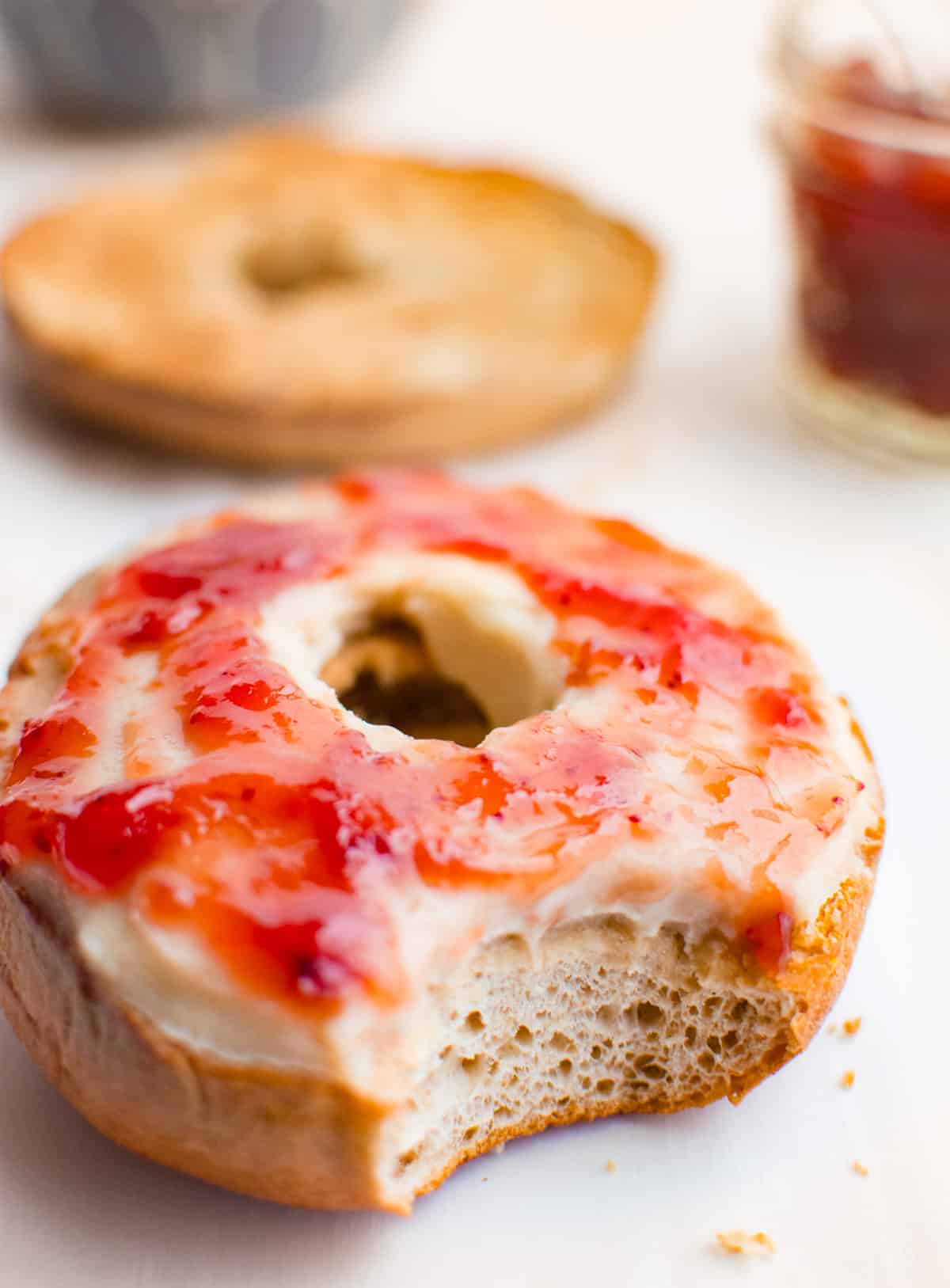 A bite taken out of a toasted bagel spread with cashew cream cheese and strawberry preserves. There is a jar of preserves and other half of toasted bagel in the background.