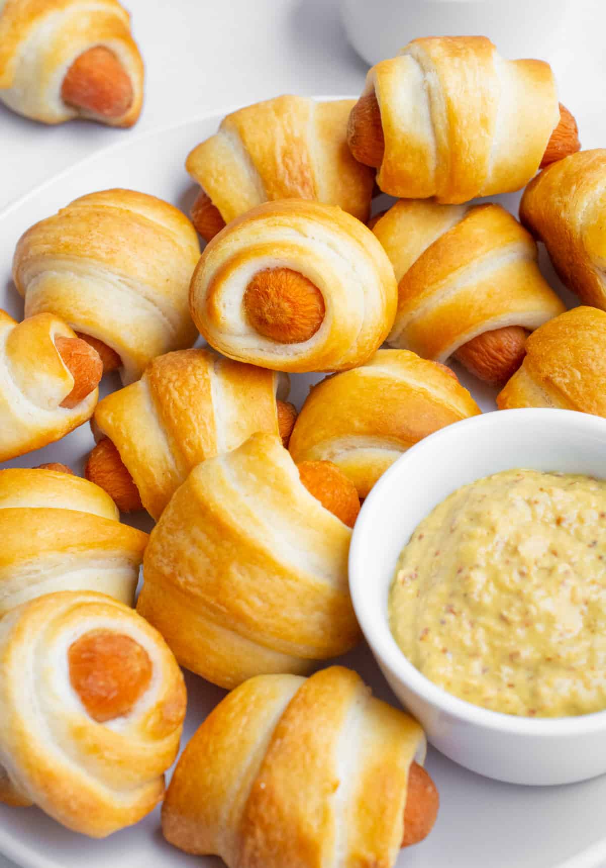 A pile of mini vegan pigs in a blanket next to a white dish of mustard dip.