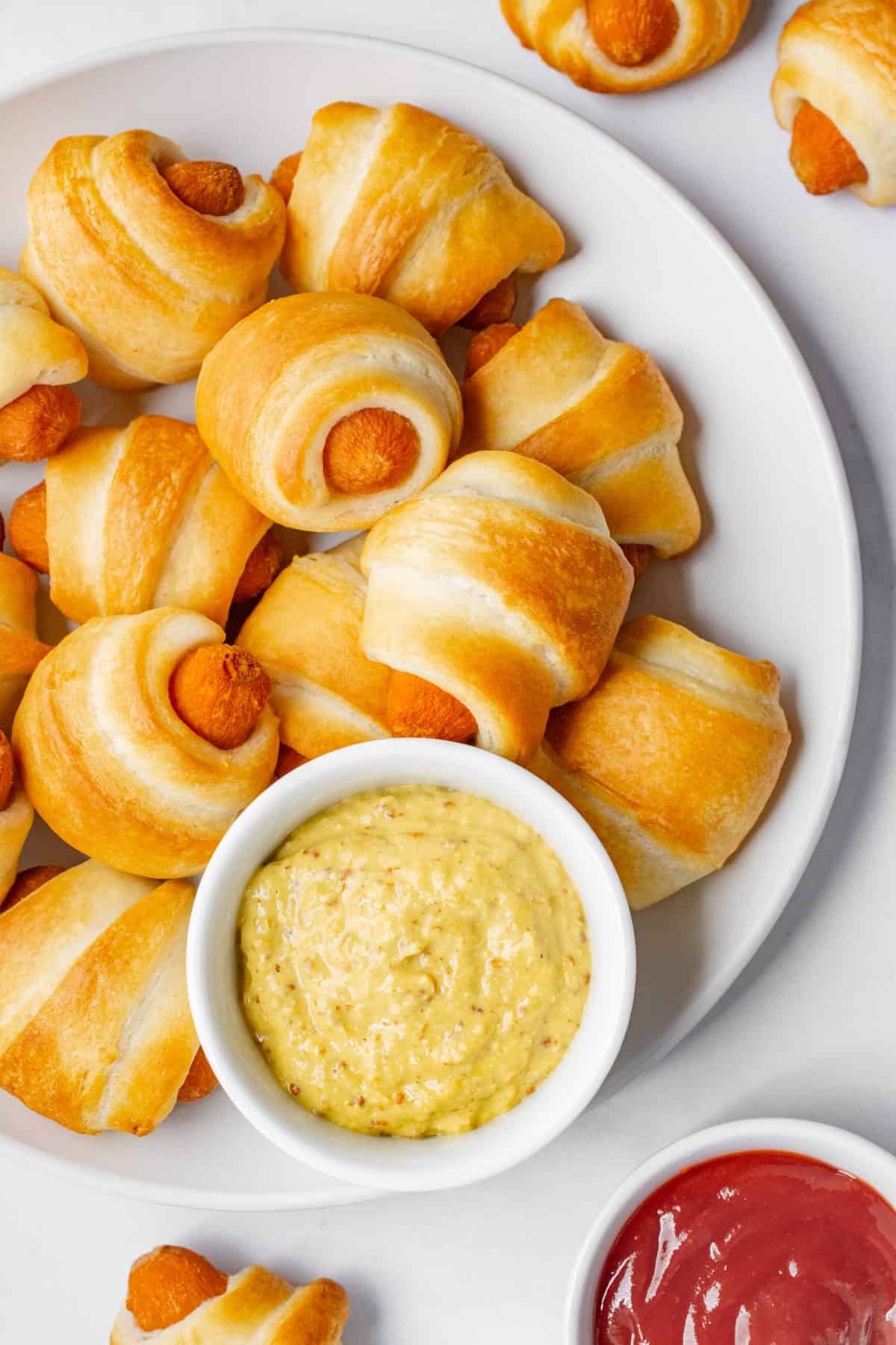 A white plate piled with vegan pigs in a blanket and a white dish of mustard dip.