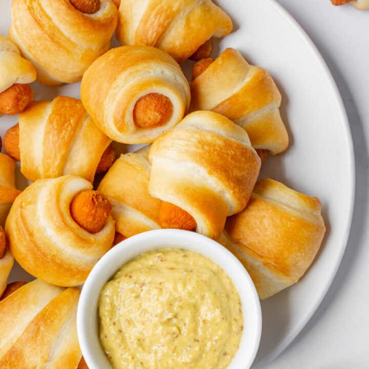 Vegan pigs in a blanket on a white plate with a small white bowl of stoneground mustard.