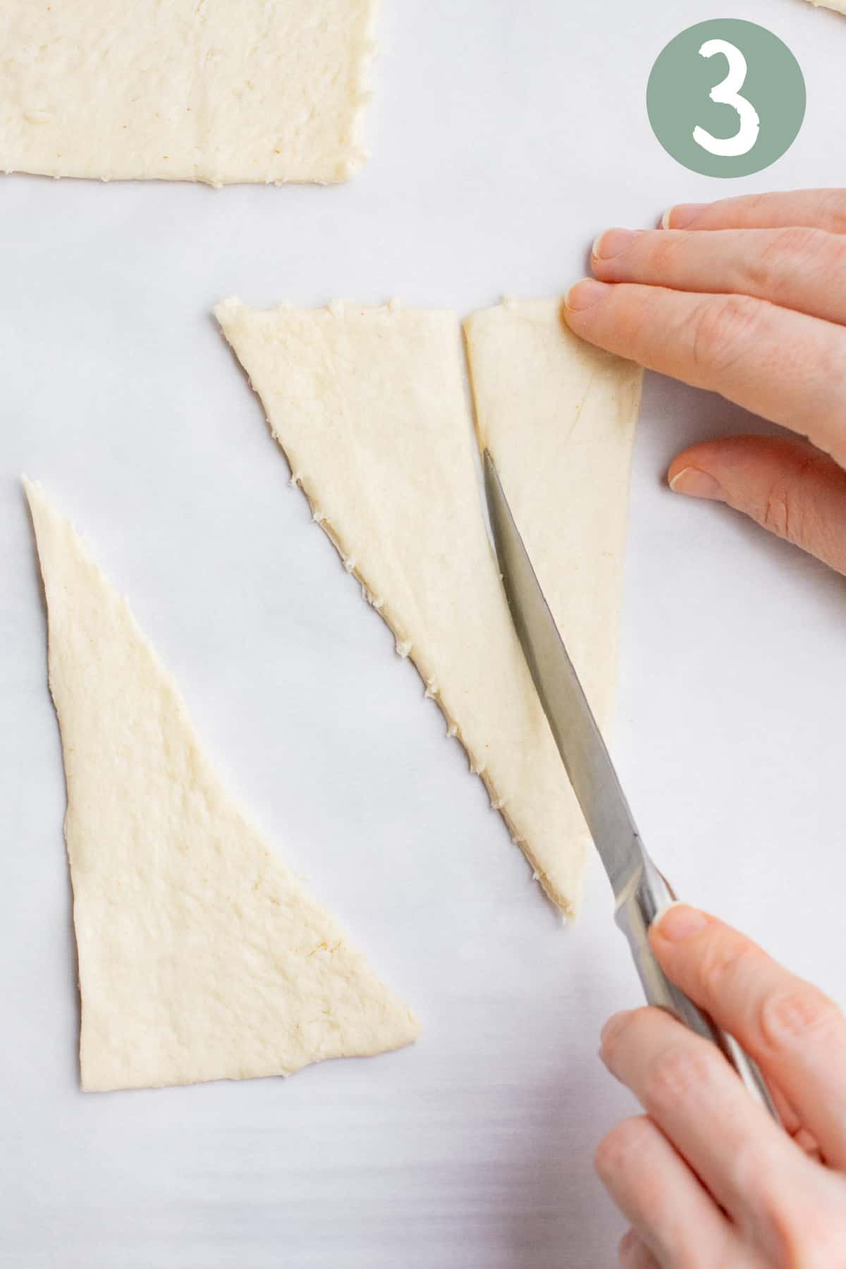 A hand holding a knife cutting a crescent roll triangle dough in half lengthwise. 