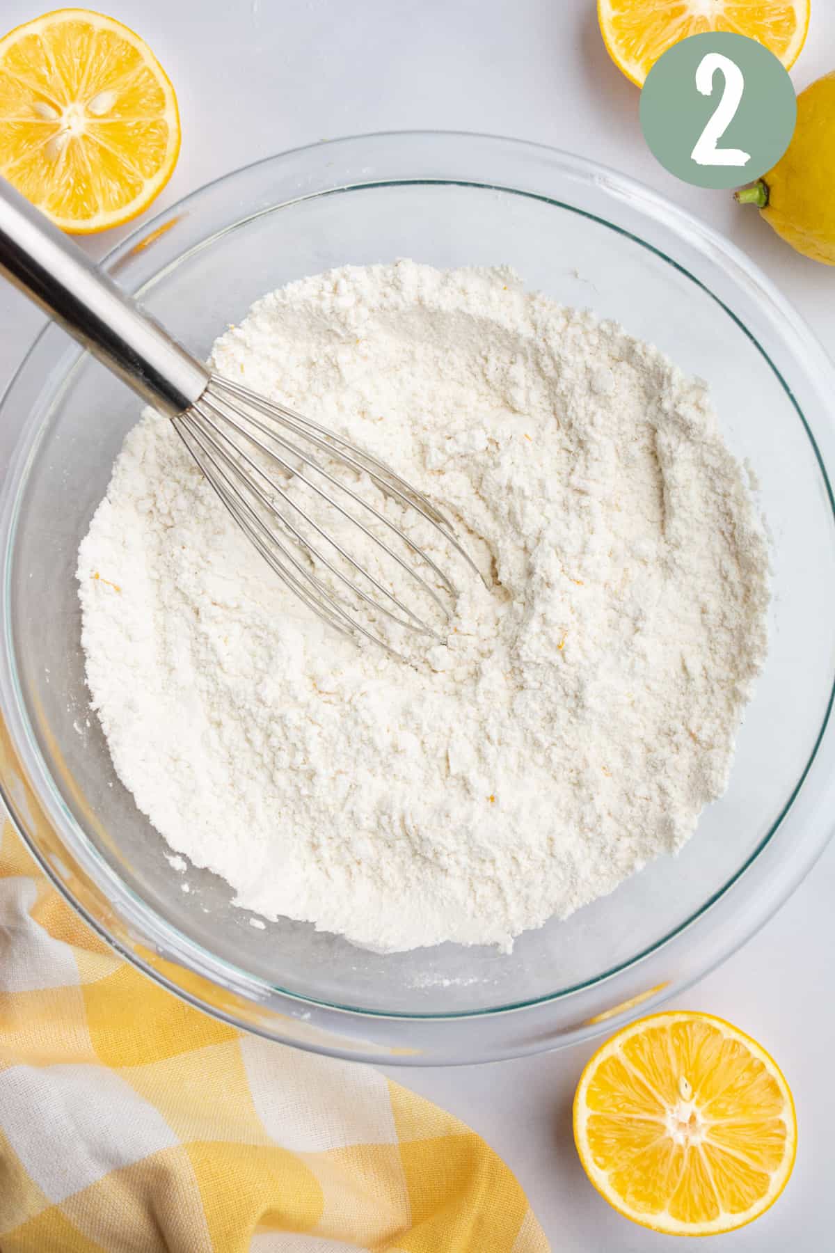 A whisk in a glass bowl of flour with cut lemon halves and a yellow checkered towel around the bowl.
