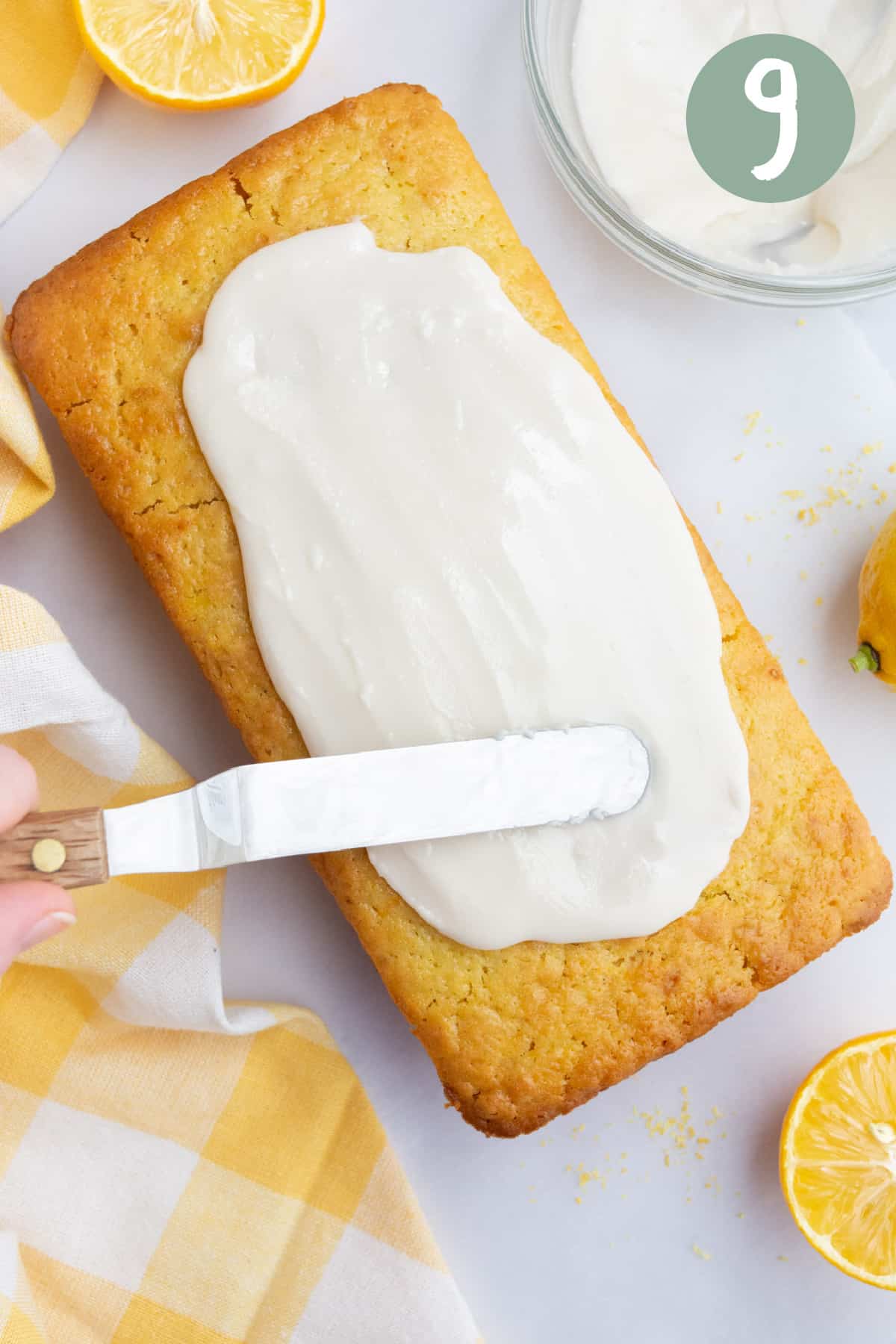 A spatula spreading icing over a vegan lemon loaf.  There is also a glass bowl of icing and a few cut lemons around the loaf.
