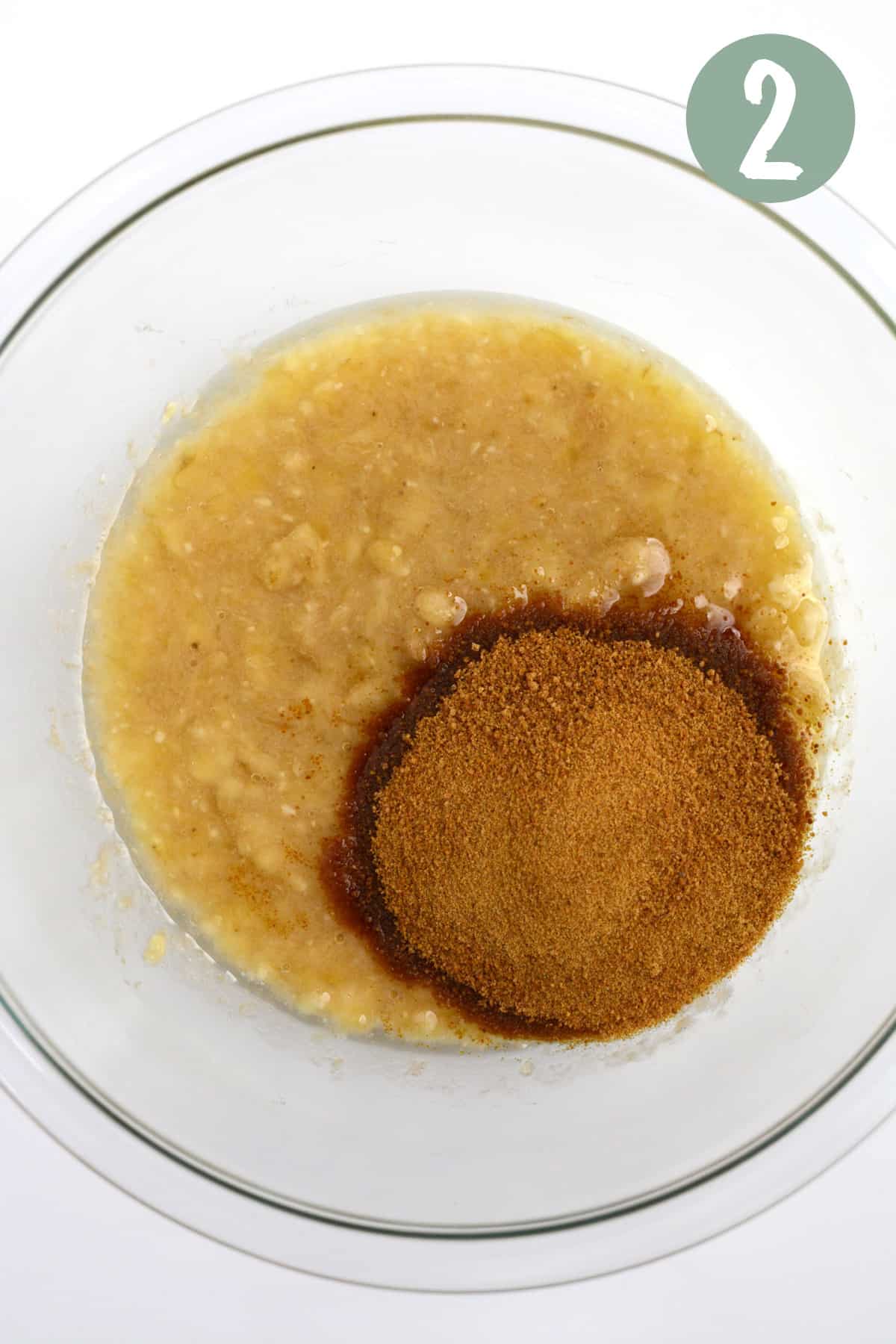 Mashed bananas and oil mixed together with a small pile of coconut sugar on top in a glass bowl.