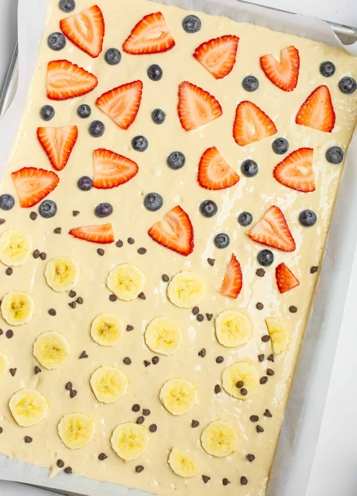 Pancake batter on a sheet pan topped with half strawberries and blueberries and the other half banana slice and chocolate chips.