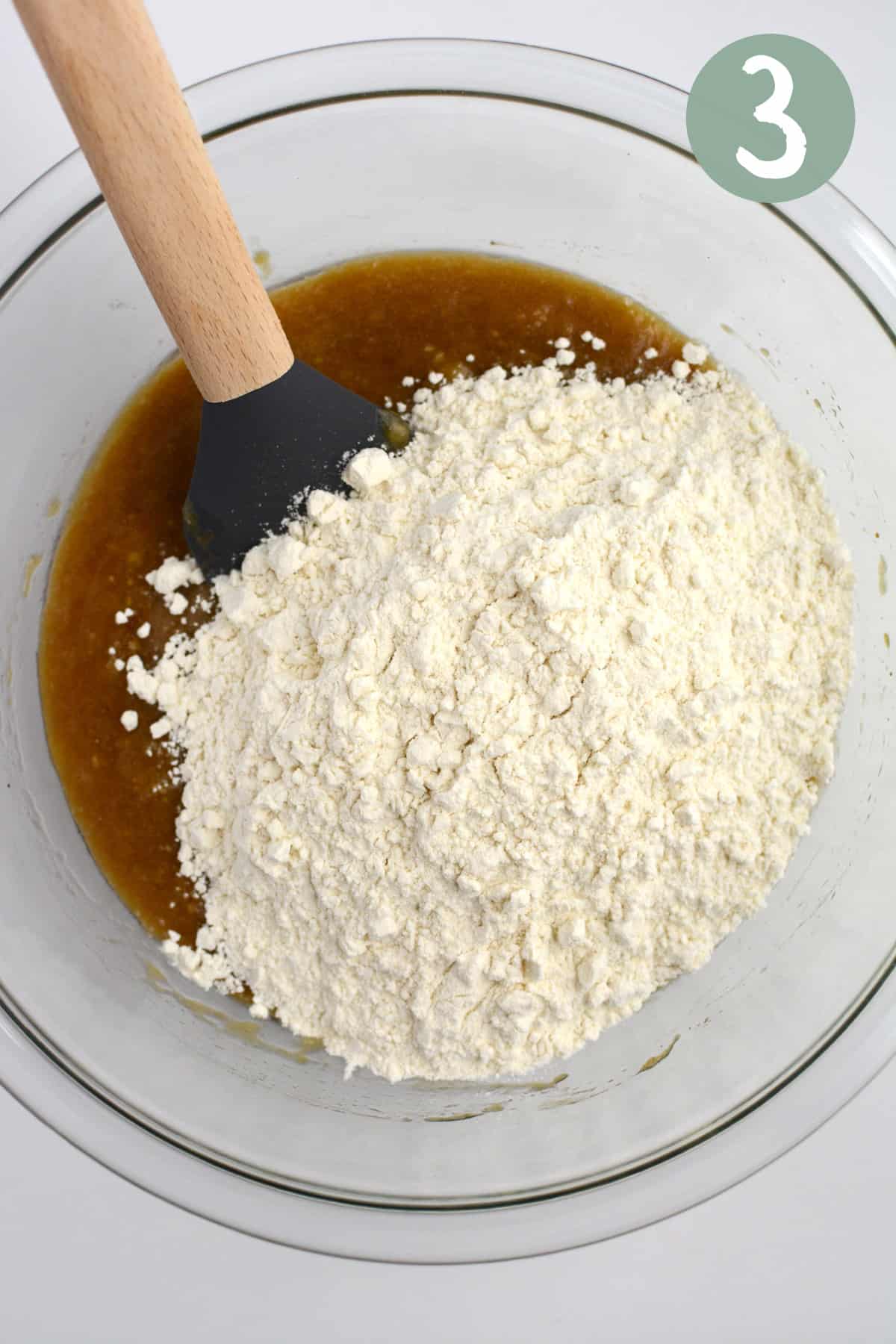 A glass bowl with mashed bananas, coconut oil, and coconut sugar mixed together and flour added on top.