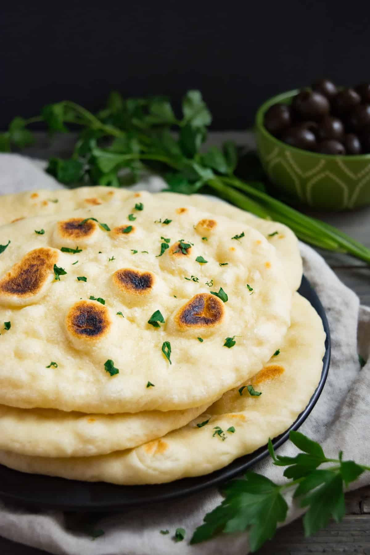 Fresh vegan naan on a black plate with olives and parsley in the background.