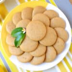 A batch of lemon cookies stacked on a plate garnished with lemon leaves and a flower.