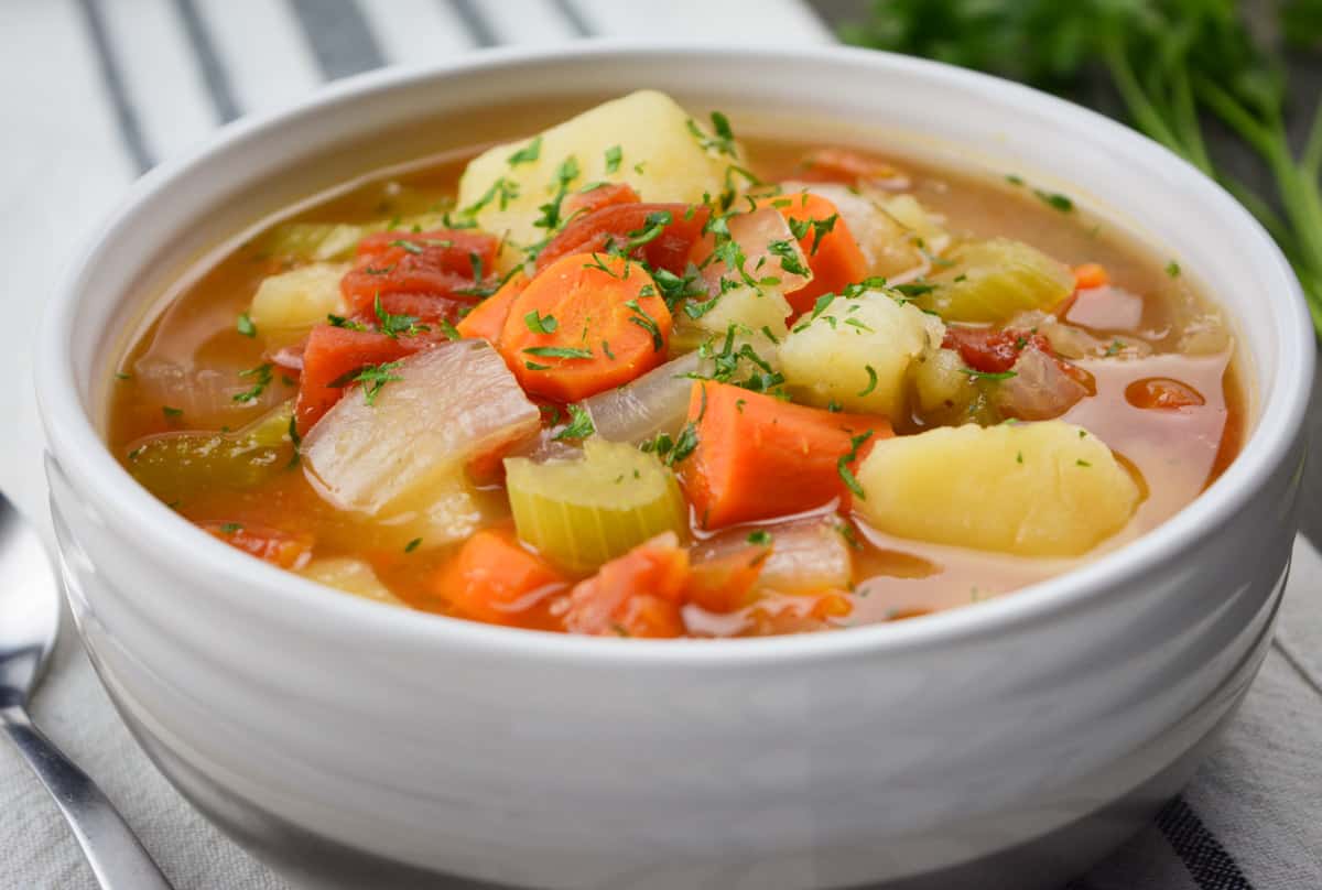 A white bowl of vegetable soup filled with carrots, celery, tomatoes, onion and potatoes. 