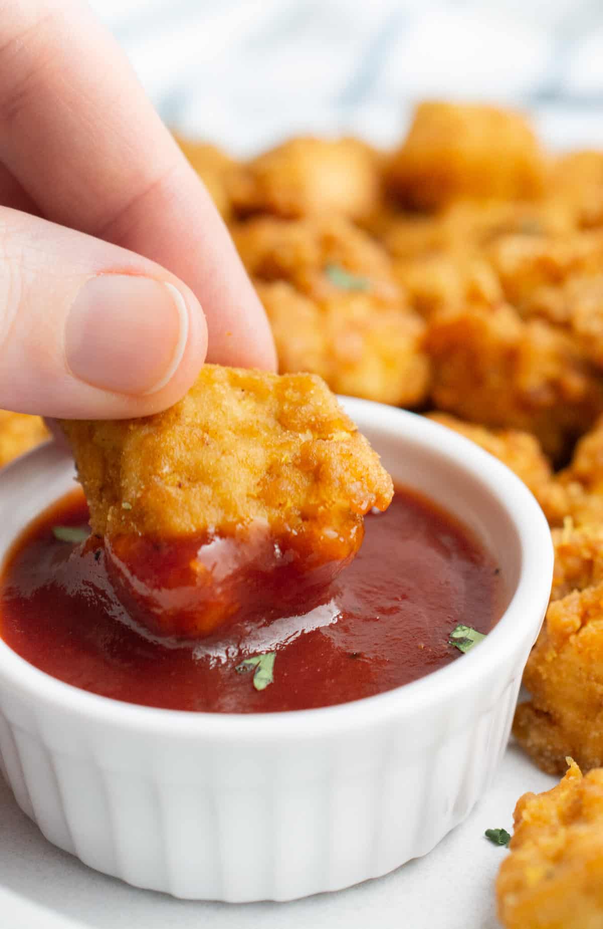 A hand dipping a tofu nugget into a bowl of BBQ sauce.