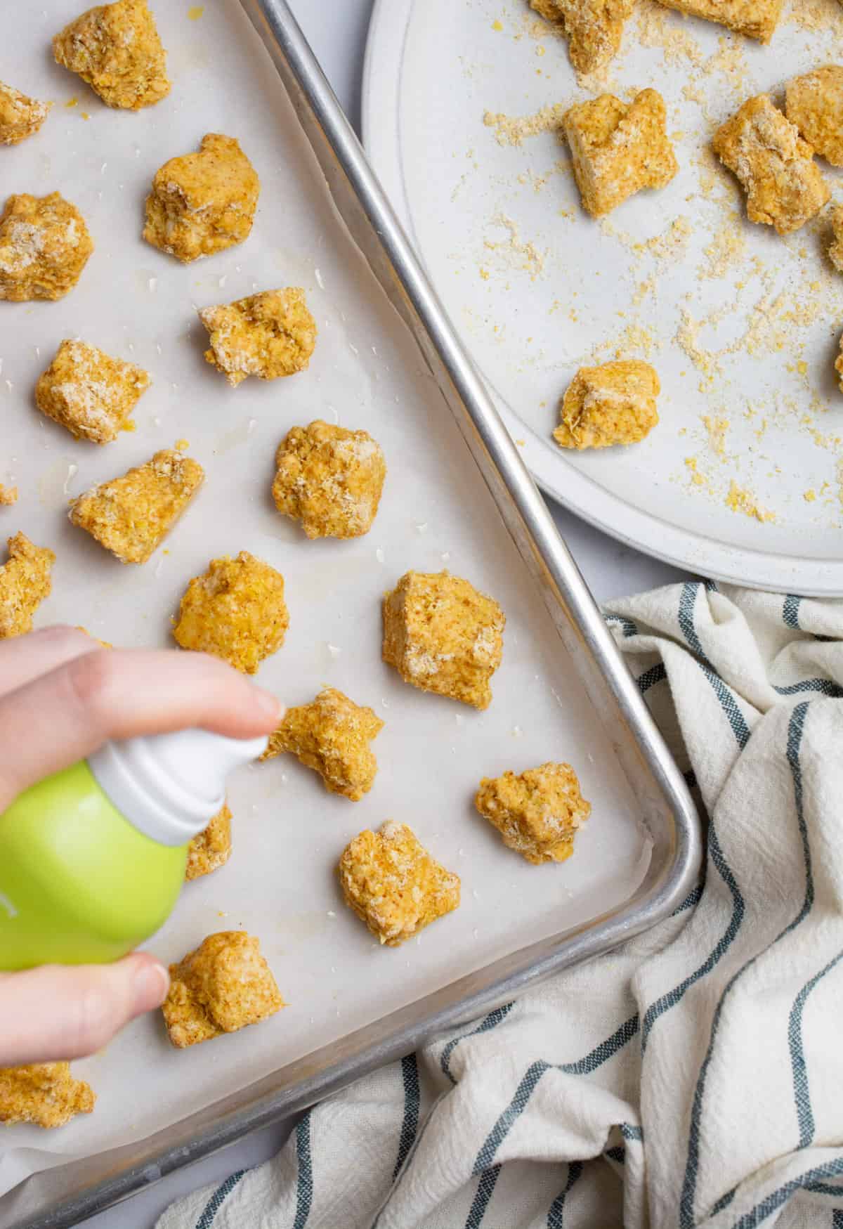 Breaded tofu nuggets on a baking sheet being sprayed with cooking oil.