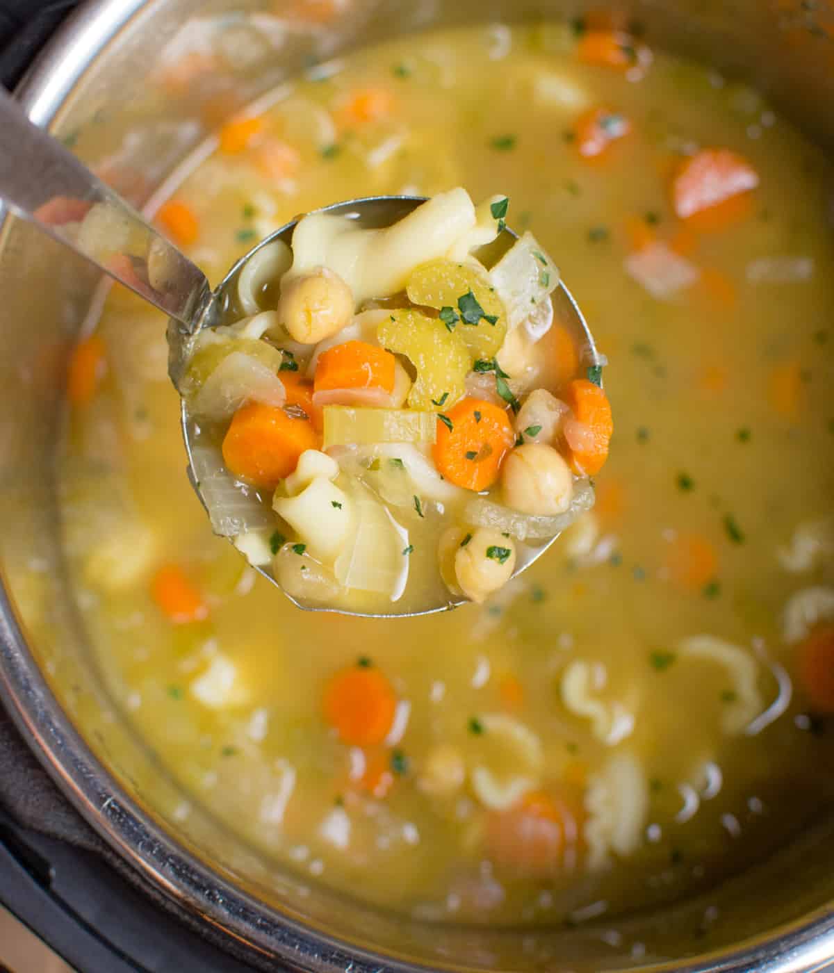 A ladle of soup filled with chickpeas, pasta, carrots, onions, and celery held over an Instant Pot full of soup.