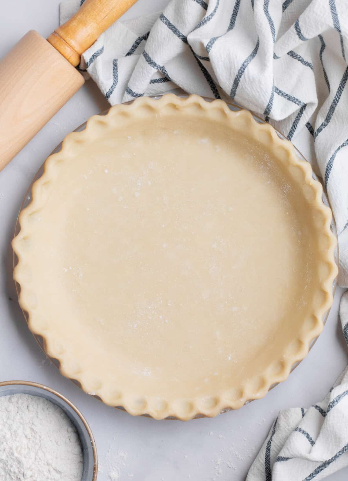 Vegan pie crust dough with crimped edges in a glass pie dish with a small bowl of flour, a rolling pin and a kitchen towel nearby.