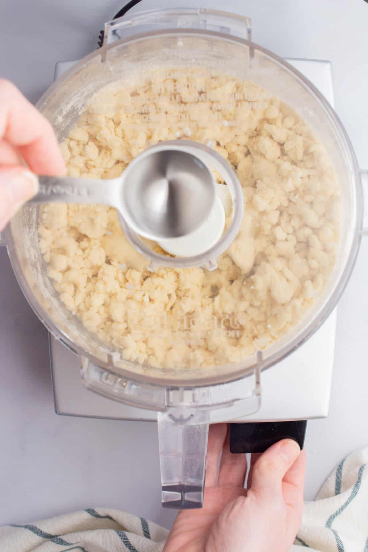 A tablespoon of water being poured into a food processor to make pie crust.