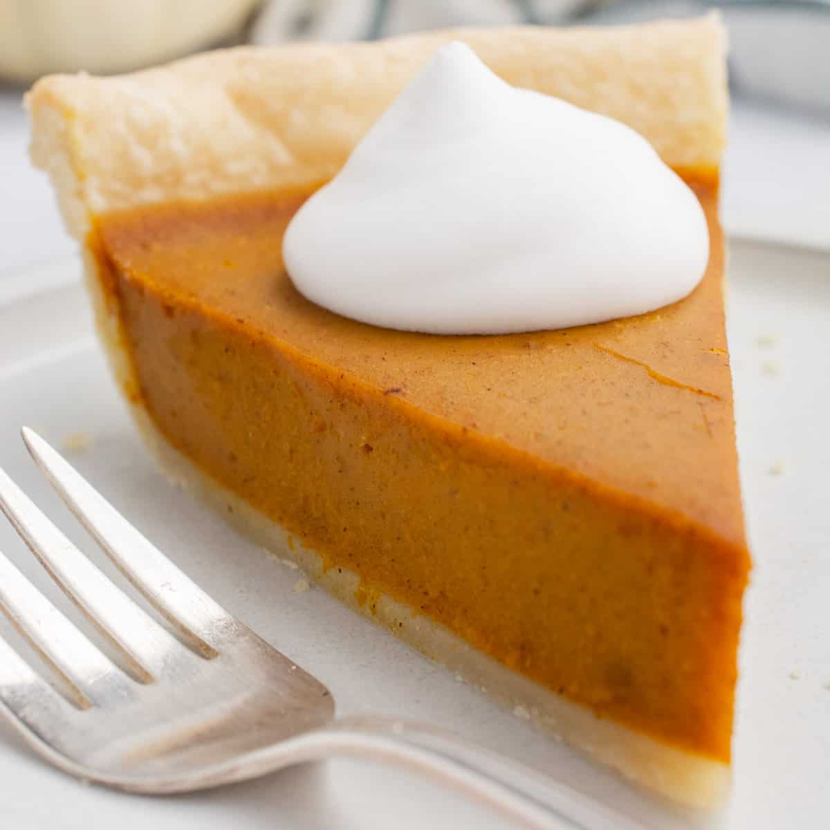 A slice of vegan pumpkin pie topped with vegan whipped cream on a white plate with a fork.
