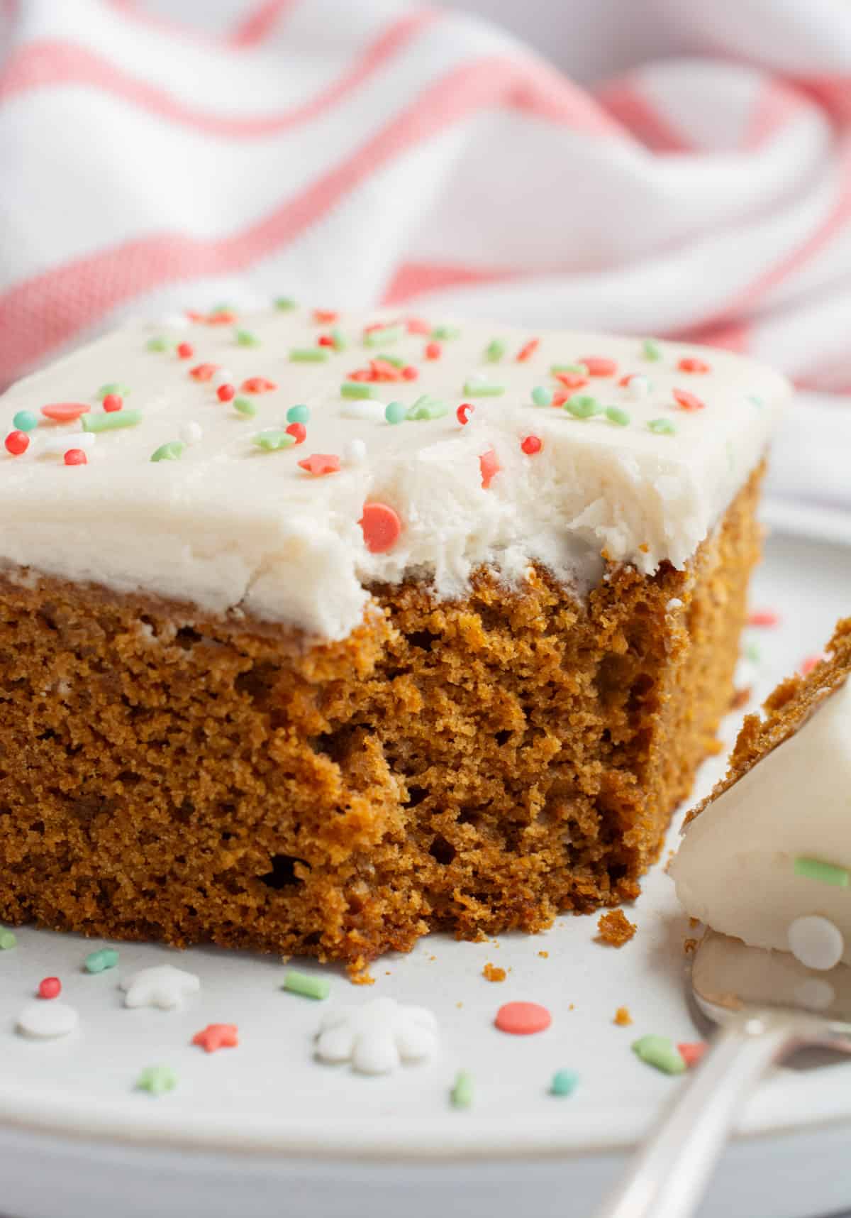 A slice of vegan gingerbread cake topped with frosting and festive Christmas sprinkles with a piece taken out with af fork.