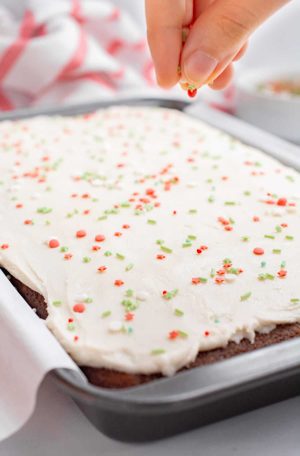 A hand sprinkling Christmas sprinkles on a frosted vegan gingerbread cake,