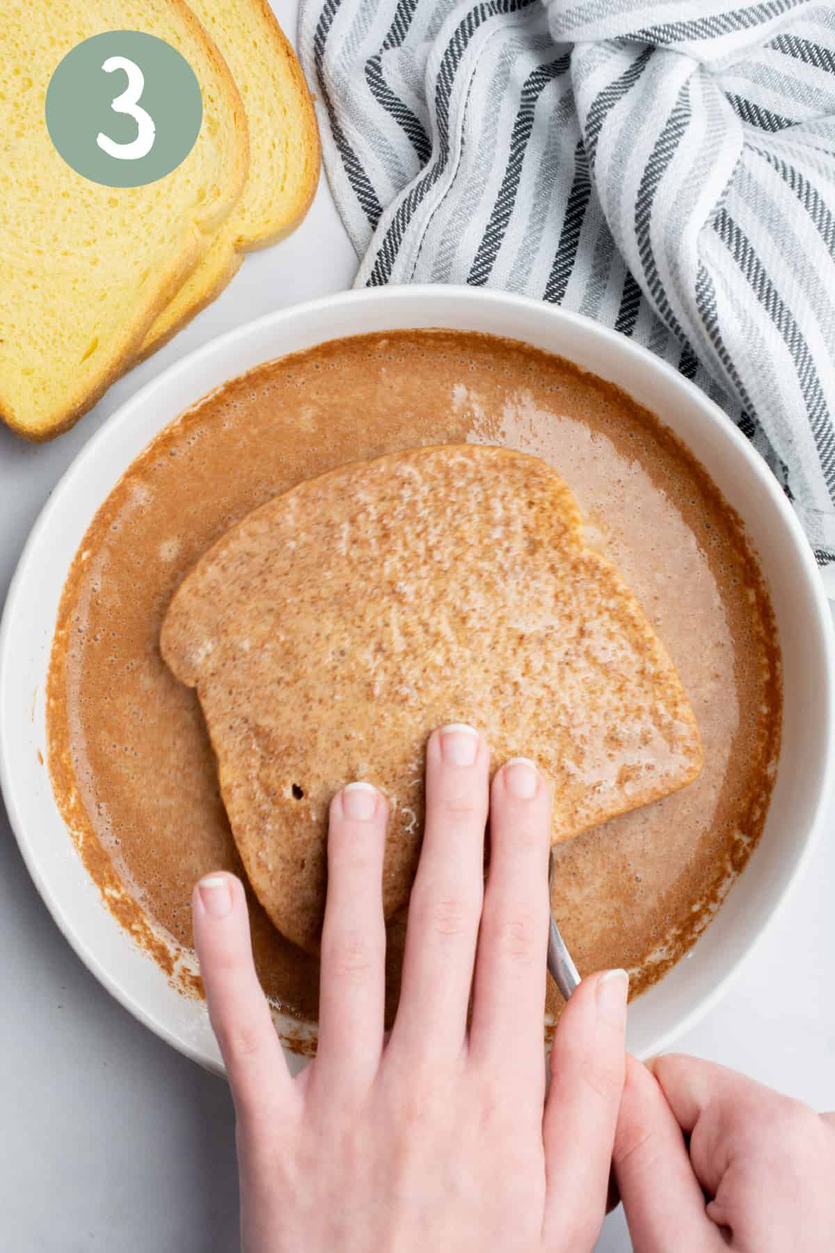 A slice of bread on a fork with a hand dipping it in a bowl of batter.