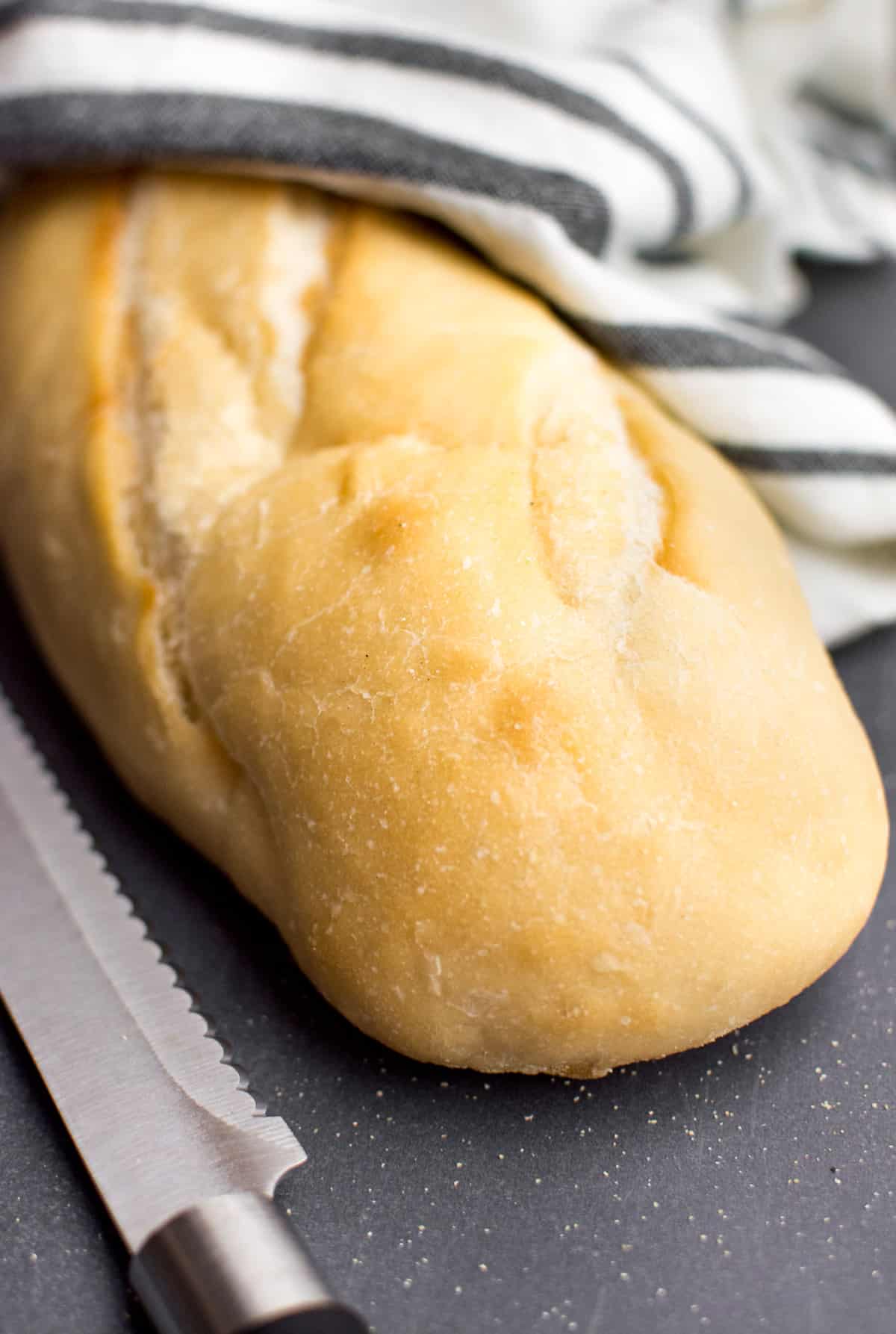 A loaf of french bread on a dark surface with a white and gray striped towel and bread knife.