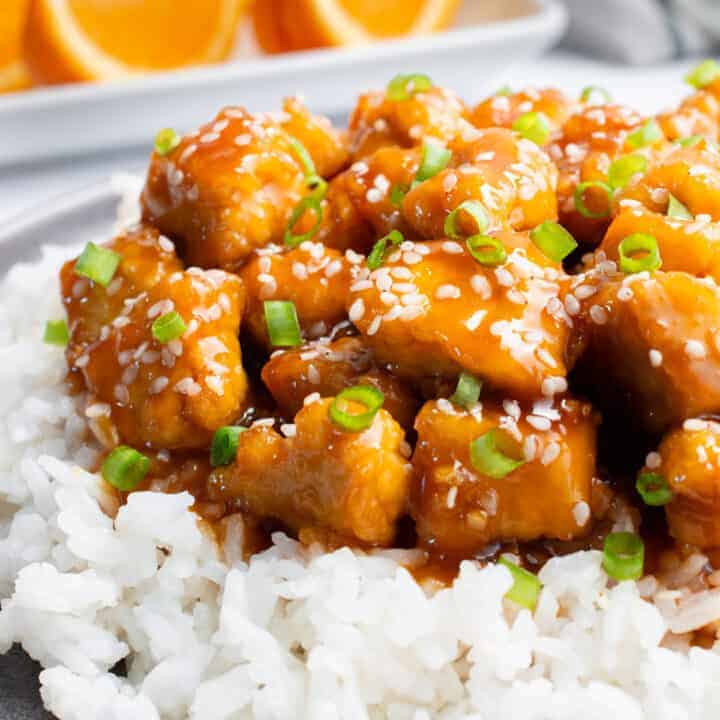 Orange tofu on a bed of white rice topped with sesame seeds and chopped green onion. There are orange slices on a plate in the background.