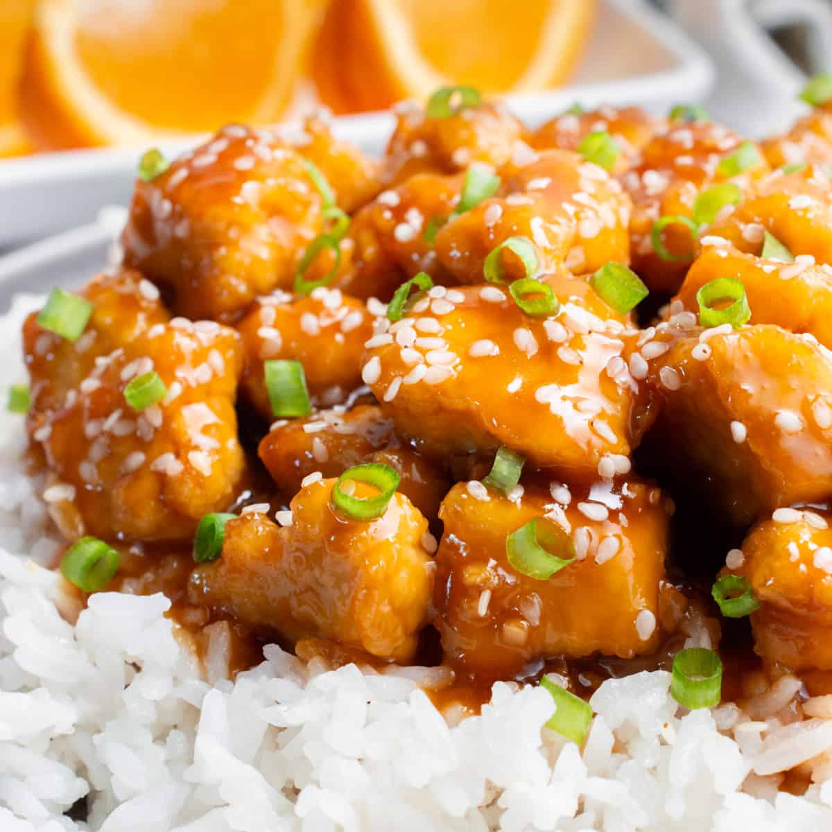 Orange tofu on a bed of white rice topped with sesame seeds and chopped green onion. There are orange slices on a plate in the background.