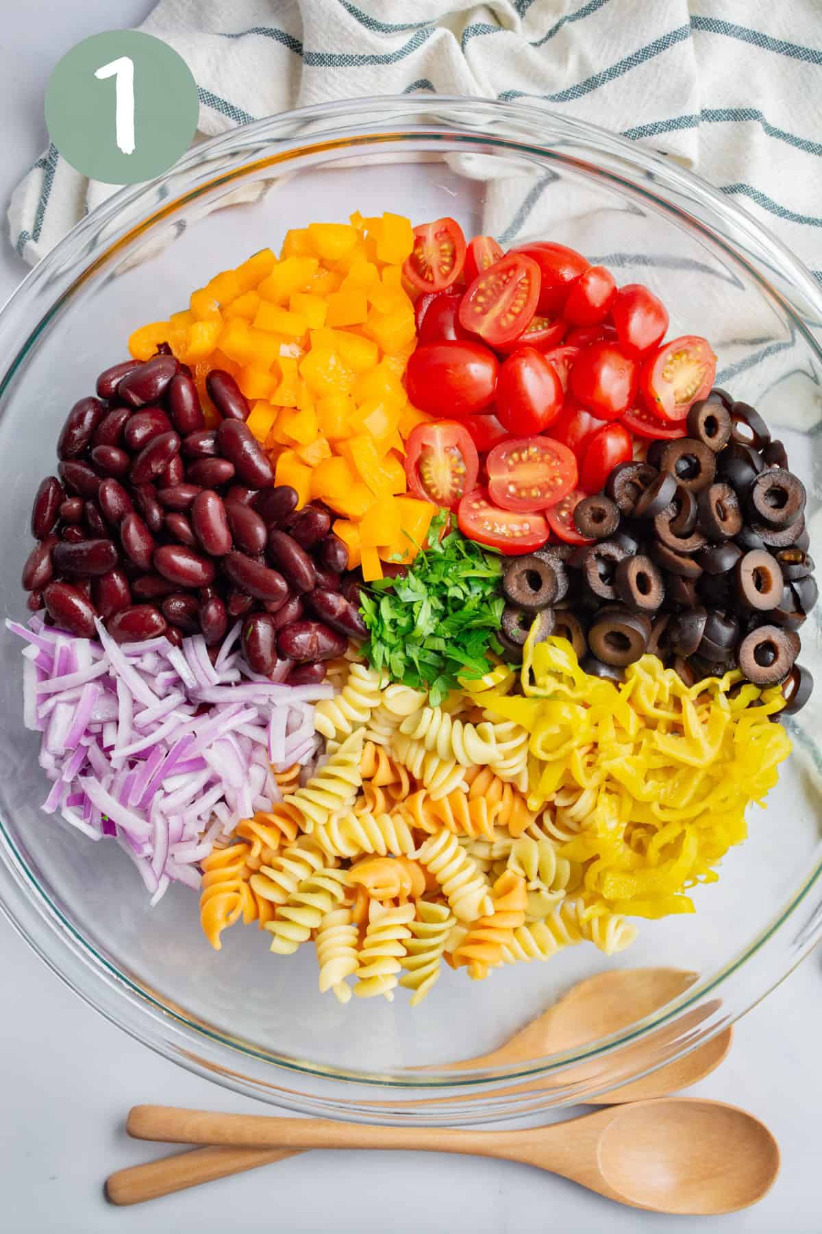 A large glass bowl with cooked spiral pasta, chopped red onion, sliced pepperoncini, sliced olives, red kidney beans, chopped orange bell pepper, and cherry tomatoes halved.