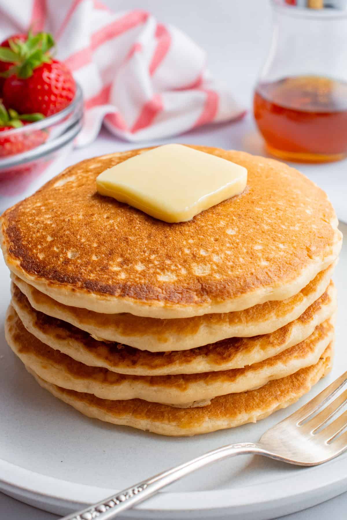 A stack of pancakes topped with a square of butter on a white plate next to a fork.