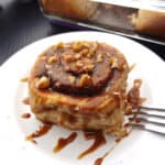 A cinnamon roll topped with maple glaze and walnuts on a white plate.