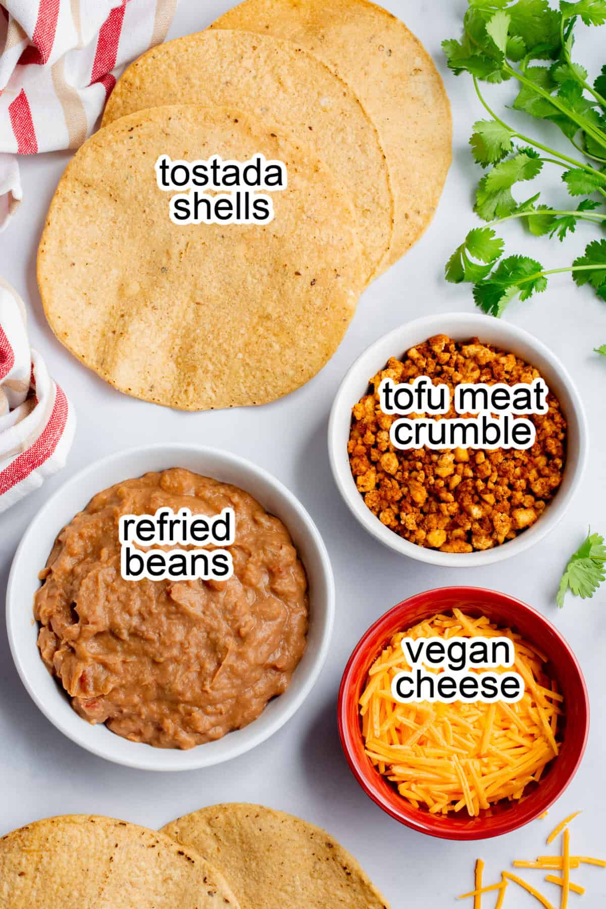 Tostada shells, fresh cilantro, refried beans in a bowl, vegan tofu meat in a bowl, and vegan cheese in a red bowl.