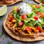 Tostada shell topped with refried beans, tofu ground beef, vegan cheese, shredded lettuce, diced tomatoes, and dairy-free sour cream.