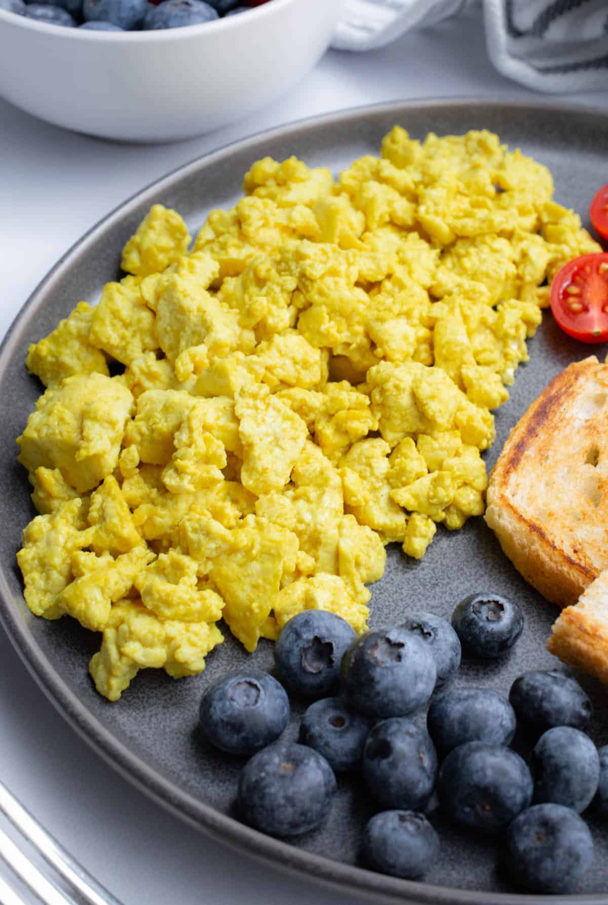 Close up of tofu on a plate with cut cherry tomatoes, toast, and blueberries.
