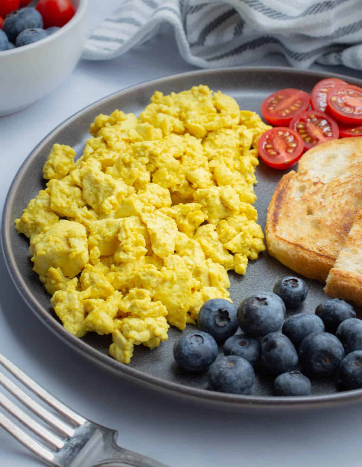 Tofu scramble on a plate with cut cherry tomatoes, blueberries, and a slice of toast.