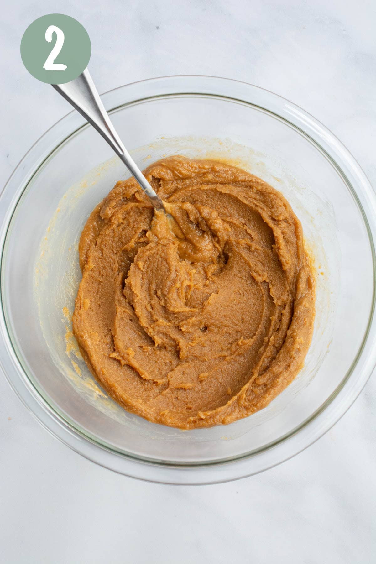 Peanut butter, sugar, and almond milk are mixed in a glass bowl with a spoon.