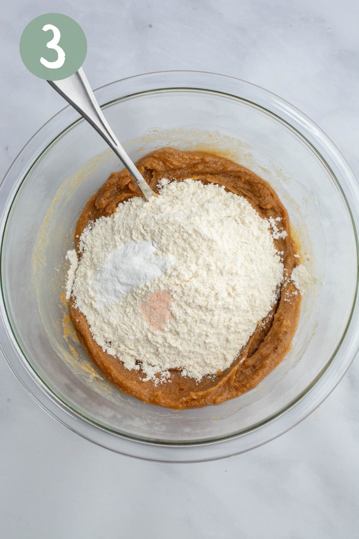 A peanut butter mixture with sifted flour, baking soda, and salt on top in a glass bowl.