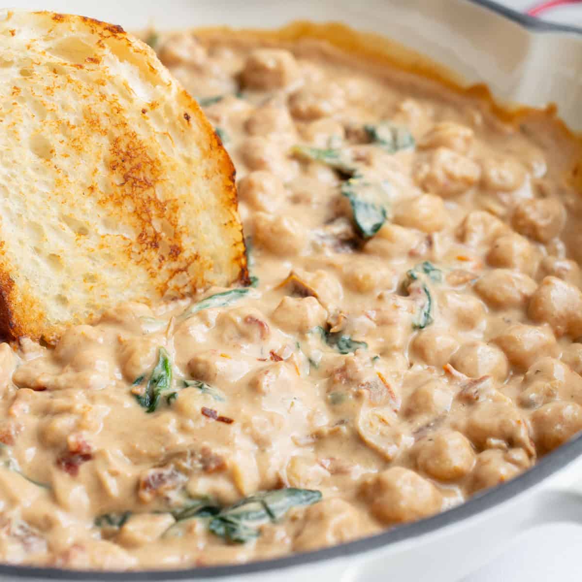 A pan of chickpeas in Tuscan sauce with a toasted slice of bread being dipped into it.