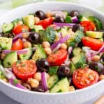 Chopped cucumber, tomatoes, red onion, olives, and chickpeas in a white bowl mixed with a olive oil and lemon dressing.