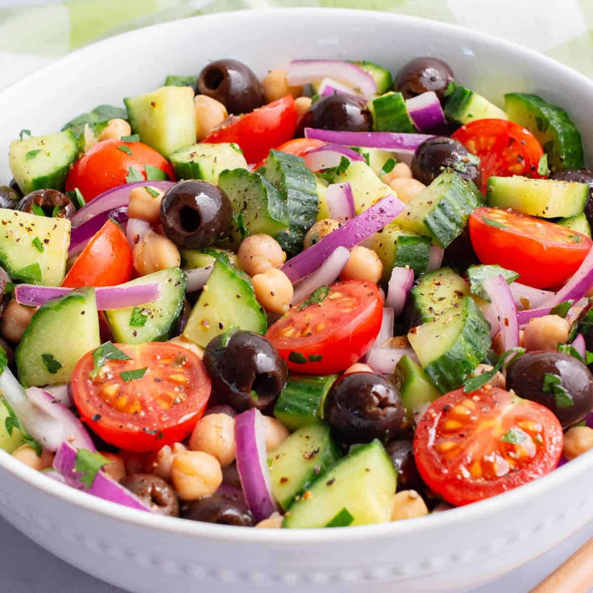 Chopped cucumber, tomatoes, red onion, olives, and chickpeas in a white bowl mixed with a olive oil and lemon dressing.