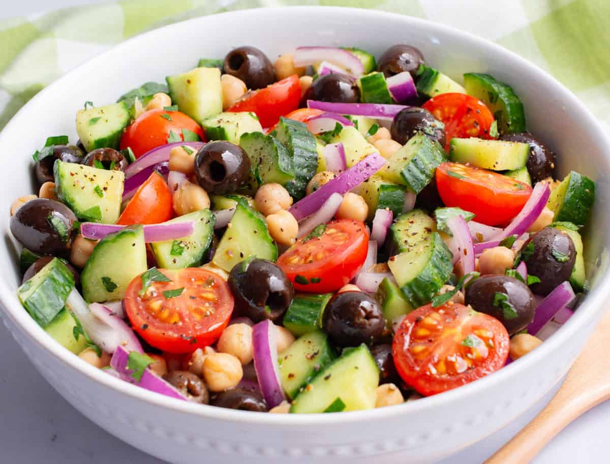 Chopped cucumber, tomatoes, red onion, olives, and chickpeas in a white serving bowl.
